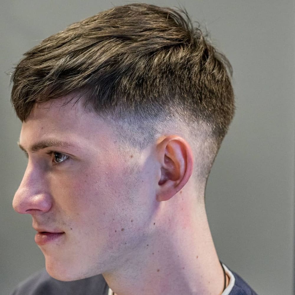 Young man with a Caesar haircut featuring a short, straight fringe and a mid fade on the sides and back. The hair on top is slightly textured, adding volume and dimension to the overall look. He is wearing a dark-colored shirt and looking to the side, showcasing the clean and balanced style.