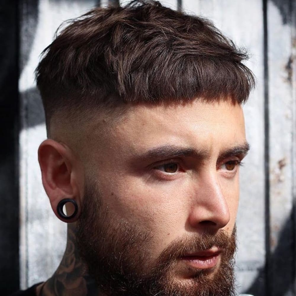 Man with a Caesar haircut featuring a short, textured fringe and a skin fade on the sides and back. He has a full beard and wears large gauge earrings, creating a bold and edgy look. The background features a weathered, rustic wall, enhancing the rugged style.