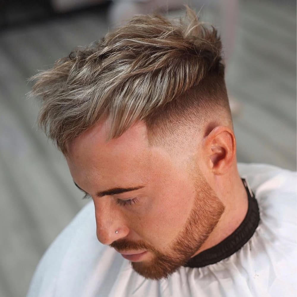 Young man with a Caesar haircut featuring a textured, tousled top and an undercut that creates a sharp contrast with the longer hair on top. The sides and back are closely shaved, highlighting the undercut. He has a neatly trimmed beard and a nose piercing, and is wearing a barber cape, seated in a barbershop with a blurred background.