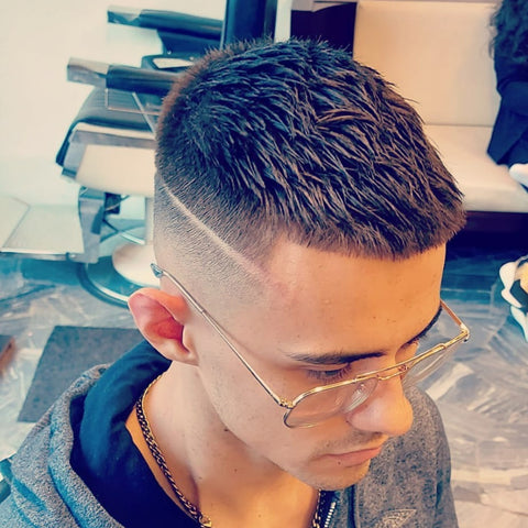 Young man with a Caesar haircut featuring a short, straight fringe and a high fade on the sides and back. The fade includes a sharp, diagonal shaved line design. He is wearing glasses, a gray hoodie, and a gold chain necklace, seated in a modern barbershop with a stylish interior.