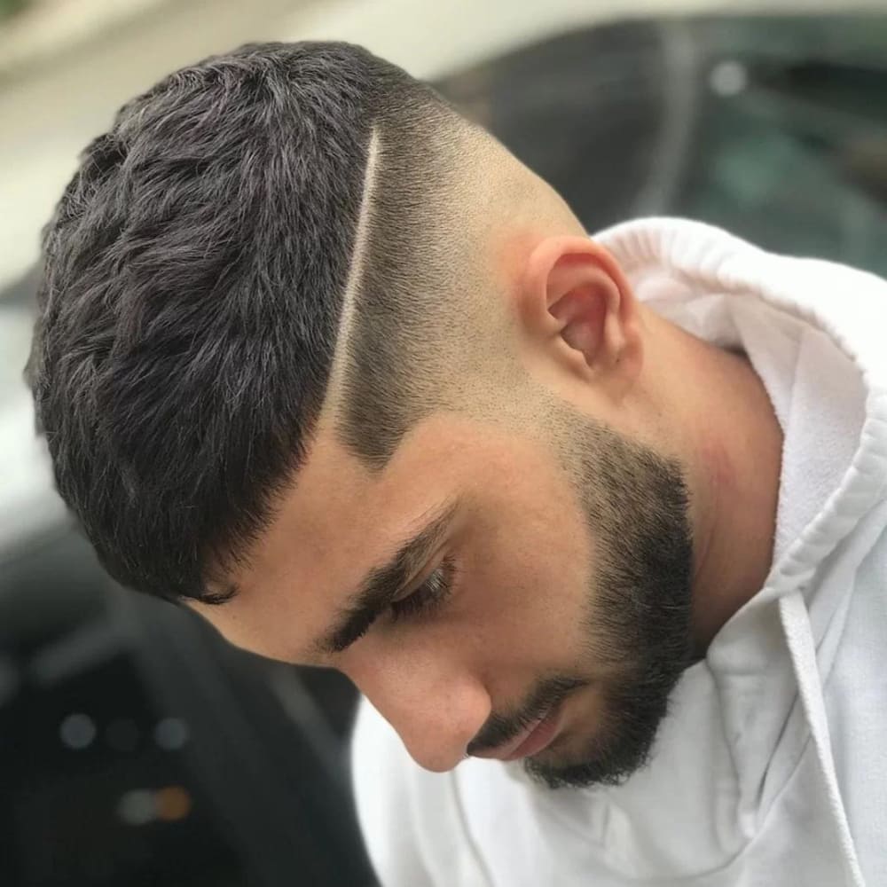 Young man with a Caesar haircut featuring a short, textured top and a prominent hard part that creates a sharp line of contrast. The sides and back have a high fade, enhancing the clean, modern look. He has a neatly trimmed beard and is wearing a white hoodie, with his head slightly tilted down, showcasing the precise haircut.