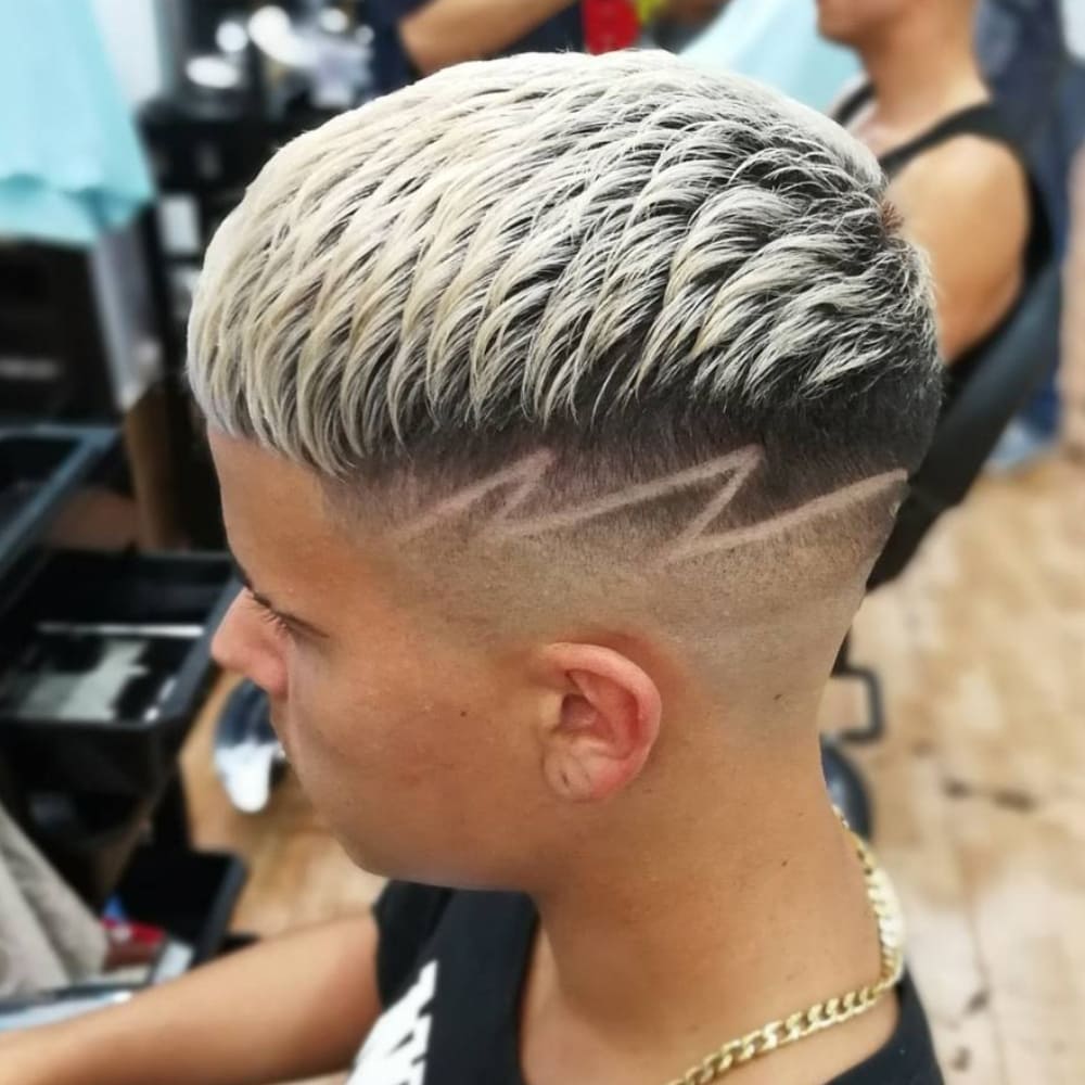 Young man with a Caesar haircut featuring bleached highlights on top, styled in a textured pattern, and a high fade on the sides and back. The fade includes an intricate zigzag shaved design, adding a bold and artistic element. He is wearing a black shirt and a gold chain necklace, seated in a busy barbershop.