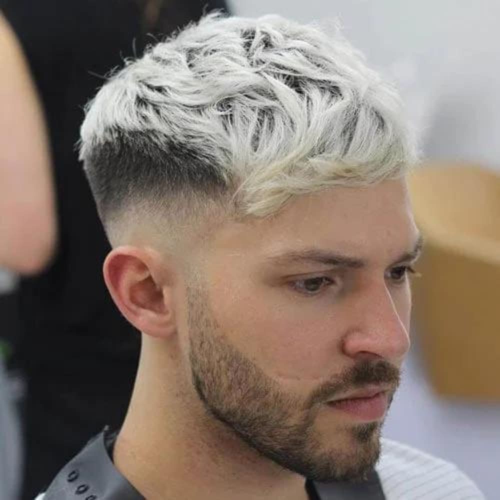 Man with a Caesar haircut featuring platinum blonde, textured hair on top with side-swept bangs. The sides and back have a high fade, creating a sharp contrast with the light-colored top. He has a well-groomed beard and is wearing a barber cape, seated in a barbershop with a blurred background.