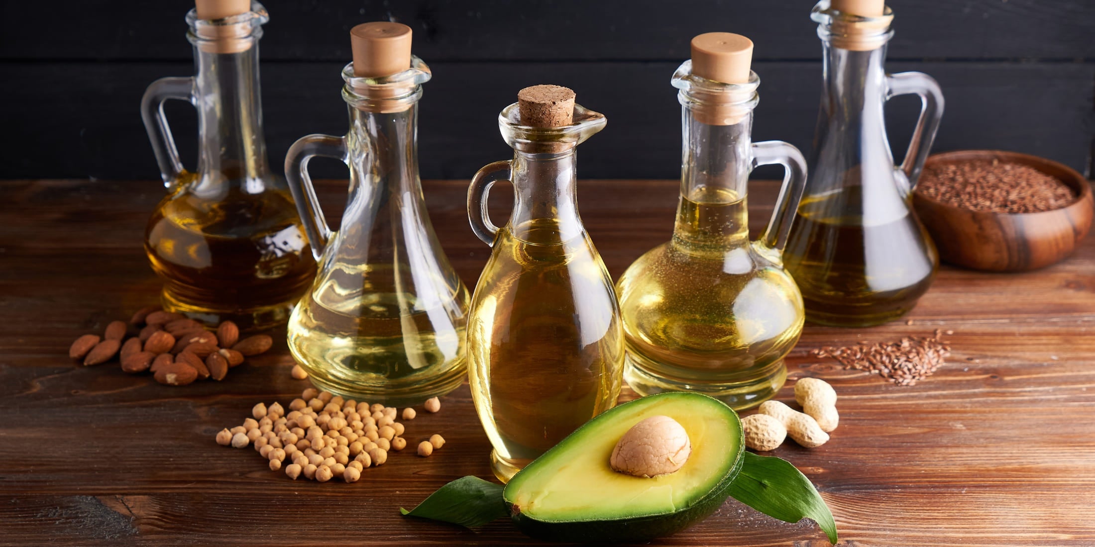 Various glass bottles filled with carrier oils, perfect for homemade shaving oil, displayed with surrounding ingredients like almonds, soybeans, and an avocado on a wooden table.