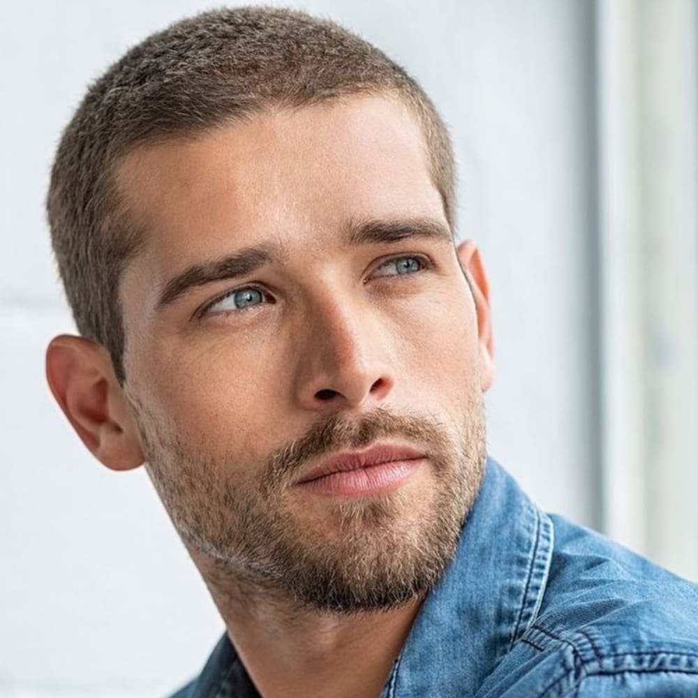 A close-up portrait of a man with a classic buzz cut hairstyle. The haircut is evenly trimmed all around, with short, uniform length hair that gives a clean and minimalistic look. The man has a neatly groomed light beard that complements the simplicity of the buzz cut, adding a touch of ruggedness to his appearance.