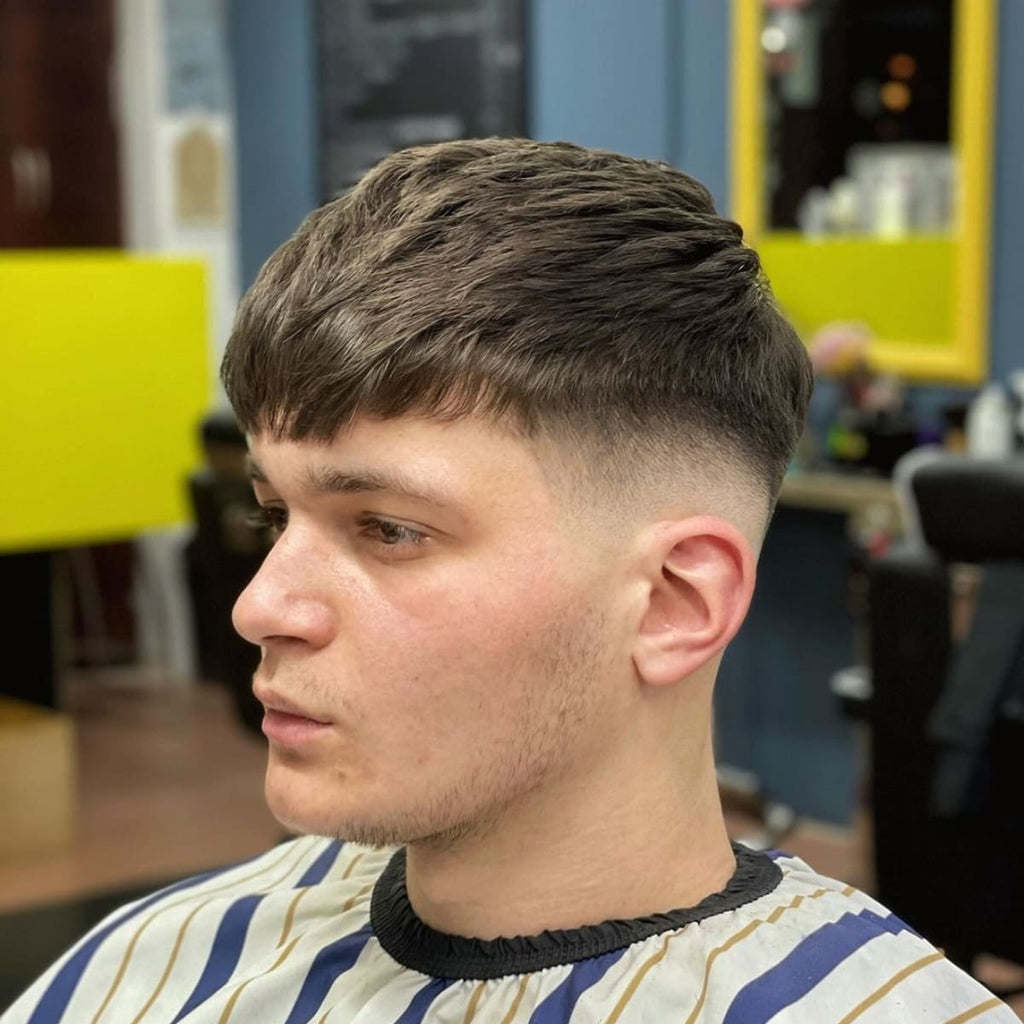 A side portrait of a young man sitting in a barber's chair, freshly styled with a short, textured haircut. The hair on top is neatly combed forward, blending smoothly into a clean fade that starts around the temples and gradually tapers down the sides and back. The man’s expression is calm and focused, while the background of the barbershop is slightly blurred, drawing attention to the crisp lines and precise cut of his hairstyle. He is wearing a barber cape with blue and yellow stripes, creating a clean and professional look.