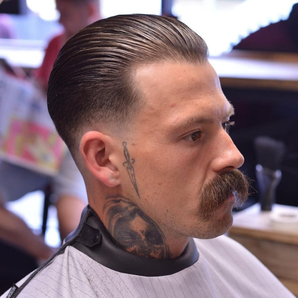 A side profile of a man in a barbershop with a classic low fade slick back haircut, featuring a sleek, combed-back top styled with a glossy finish. The low fade begins at the base of the neckline and gradually tapers just below the ears, maintaining a clean and traditional look. The man sports a bold mustache and visible neck and side tattoos, adding personality and edge to the classic hairstyle. The barbershop background includes blurred activity and natural lighting, keeping the focus on the precise details of the haircut.