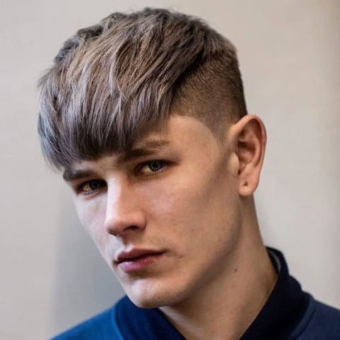 A young man is shown with a combed-down fringe hairstyle, featuring a sleek and straight fringe that falls evenly across his forehead. His hair is a cool ash-blonde shade, with subtle darker tones adding depth and dimension to the style. The top is left longer, allowing the fringe to be combed forward and down in a smooth, polished manner, while the sides are tapered and neatly faded, providing a clean and modern contrast. The man's expression is serious, with his blue eyes giving a piercing look that complements the sharpness of the haircut. He is dressed in a dark blue jacket, which adds to the cool and refined aesthetic of the overall look. The background is a soft, neutral tone, which keeps the focus on his hairstyle and facial features, highlighting the meticulous grooming and stylish appearance.