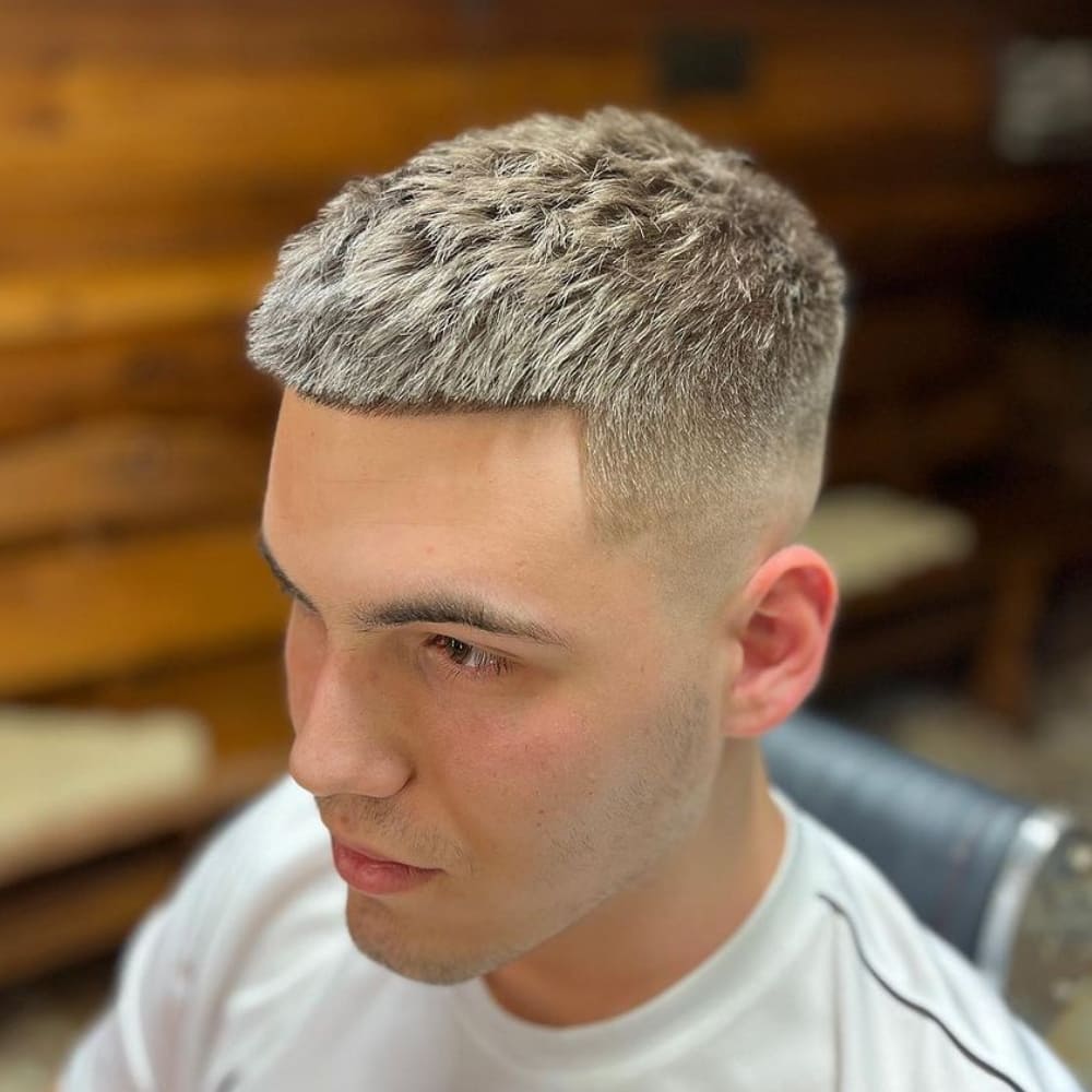 A close-up image of a young man with a crew cut buzz cut hairstyle. The haircut features short, textured hair on top with a slightly longer length, creating a rugged yet neat appearance. The sides and back are tapered into a clean fade, blending smoothly into the top. The hairline is sharply defined, with a straight line-up at the forehead, adding a crisp and polished edge to the overall look. The hair is light in color, possibly dyed, enhancing the texture and making the style stand out. The background is softly blurred, focusing attention on the sharp and modern haircut.