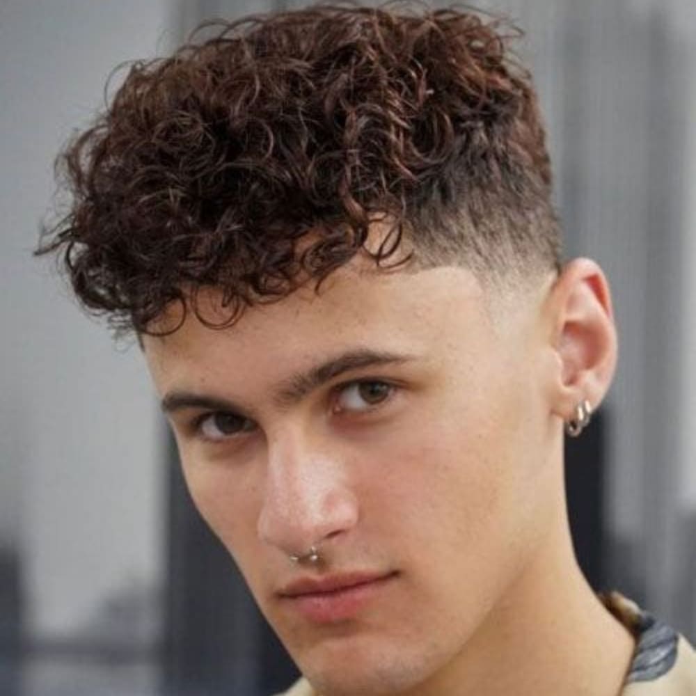 Young man with a curly Caesar haircut featuring a short, curly top and a high fade on the sides and back. The curls are natural and voluminous, adding texture and height. He has a septum piercing and wears small hoop earrings in both ears, creating a modern and stylish look. The background is blurred, focusing attention on the detailed haircut.