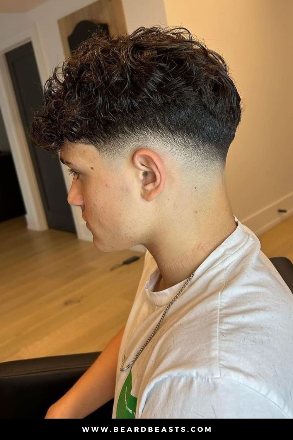 A close-up image of a young man with neatly trimmed facial hair, featuring a stylish haircut that seamlessly transitions from shorter hair on the sides to fuller, well-defined curls on top.