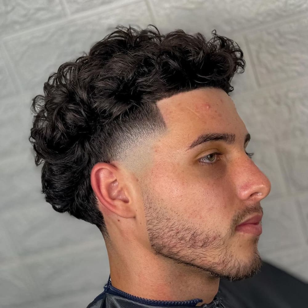 Man with a curly high taper haircut, featuring defined curls on top and sharply tapered sides. The style highlights a clean hairline and a neatly trimmed beard for a polished look.