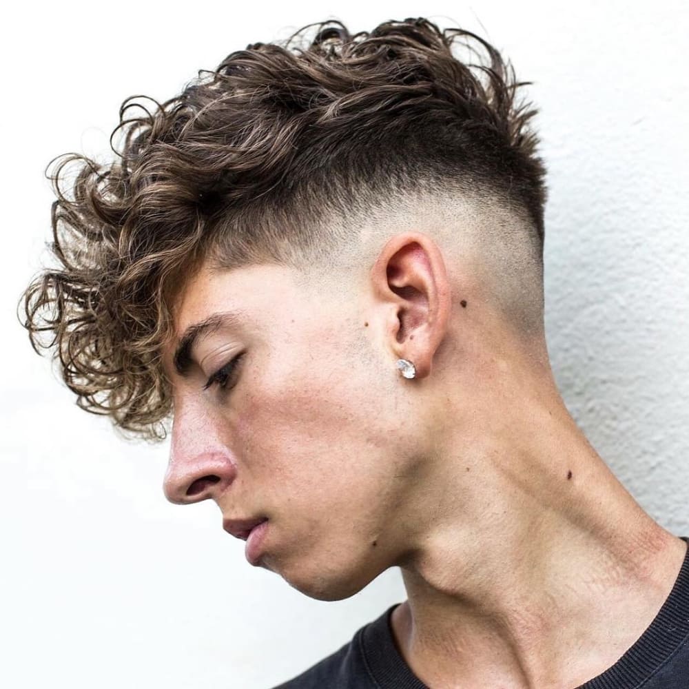 A young man with a curly textured crop haircut is depicted in the image. His hair features a voluminous and curly top, styled with natural texture and movement. The sides and back are closely faded, creating a sharp contrast with the longer curls on top. He is wearing a stud earring, which adds a touch of personal style. The overall look is modern and stylish, showcasing how a textured crop hairstyle can beautifully enhance curly hair. The setting appears to be a simple, clean background, drawing focus to his haircut and facial features.