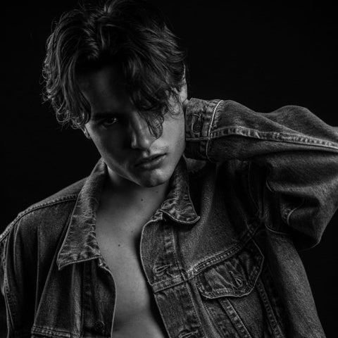 A young man is captured in a black-and-white photograph, showcasing a classic curtain bangs hairstyle. His medium-length hair is parted in the middle, with the bangs sweeping down on either side of his face, creating a soft, face-framing effect. The curtain bangs fall naturally, with some strands casually draping over his forehead, contributing to a moody and effortlessly cool look. He is wearing an oversized denim jacket, left unbuttoned to reveal his bare chest underneath, adding to the casual and edgy vibe of the image. The lighting is dramatic, with shadows emphasizing the contours of his face and the texture of his hair. His expression is intense and slightly melancholic, with his left hand casually resting on the back of his neck, further enhancing the overall moody atmosphere of the portrait. The dark background adds contrast, making the details of his hairstyle and outfit stand out prominently.