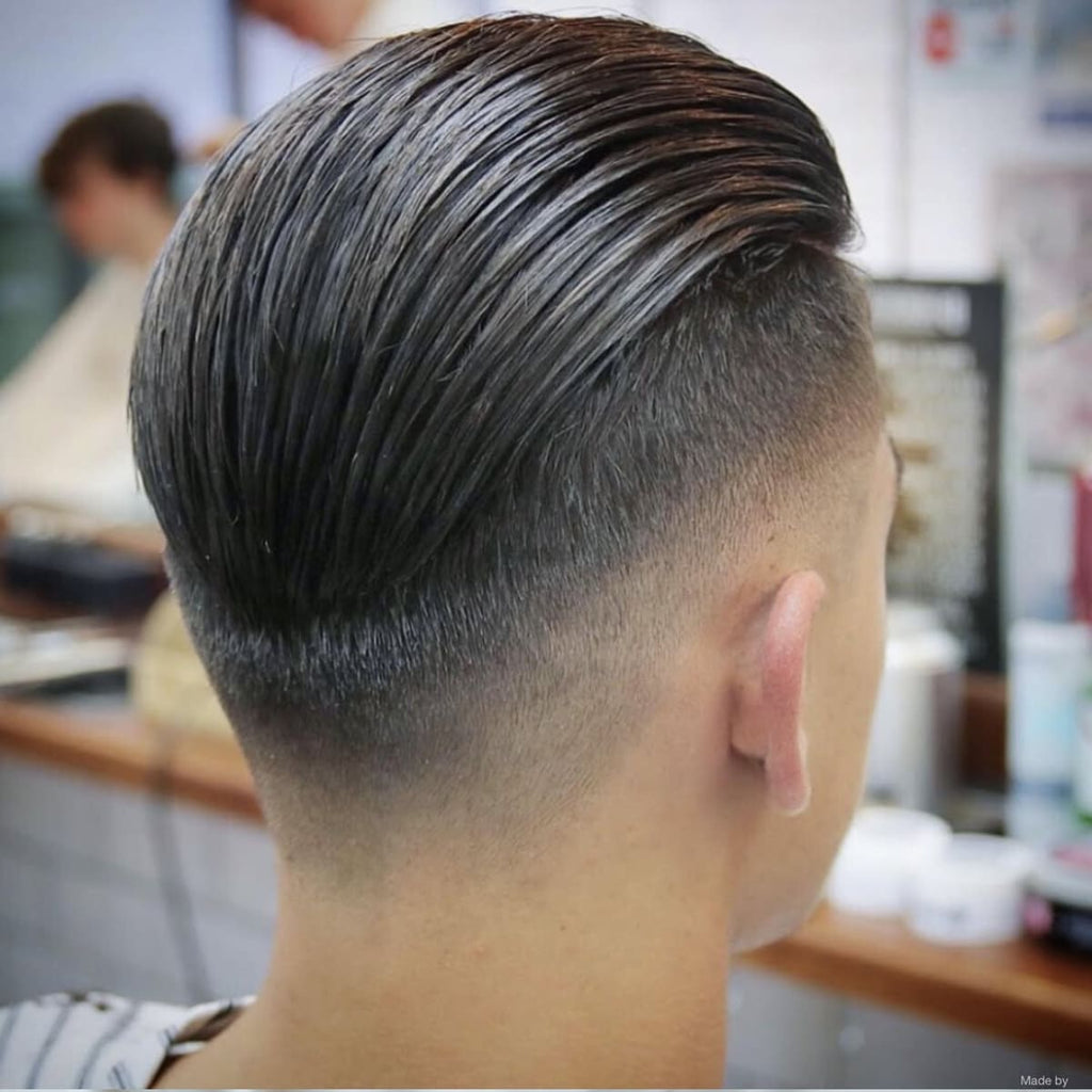 A back profile of a man with a disconnected low fade slick back haircut, showcasing a sleek and glossy top styled neatly back with a sharp, defined line separating it from the fade. The low fade begins at the base of the neckline and transitions subtly below the ears, creating a clean and gradual taper that contrasts with the longer, disconnected top. The precise styling emphasizes the sharpness of the disconnect and the smooth gradient of the fade. The background features a barbershop setting with blurred details of styling products and tools, highlighting the haircut as the focal point.