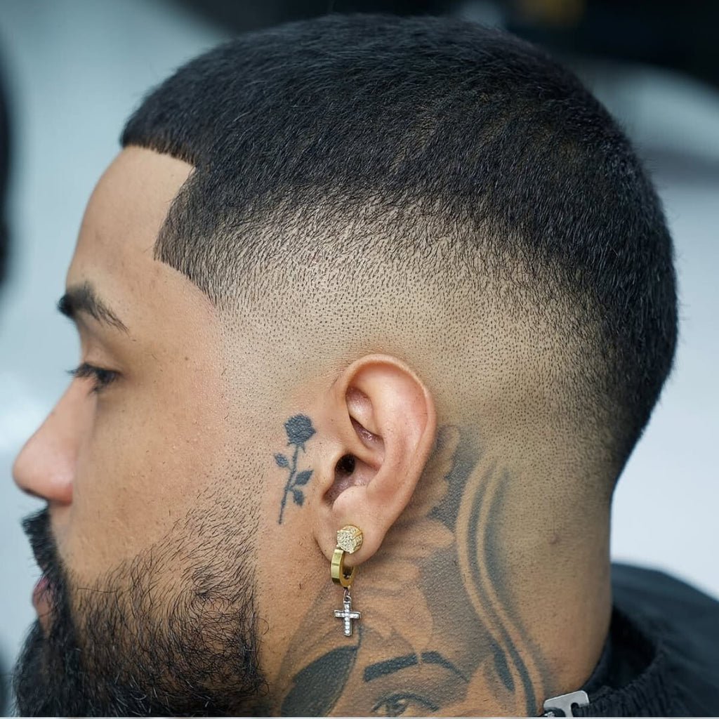 A close-up side view of a man with a skin fade buzz cut, showcasing a drop fade that curves around the ear for a sleek and modern look. The hairstyle transitions smoothly from the skin to short, textured hair on top. The style is enhanced by a full, well-groomed beard, along with visible tattoos and gold jewelry that add personality and flair. The precise lines and sharp fade highlight the clean and polished appearance.