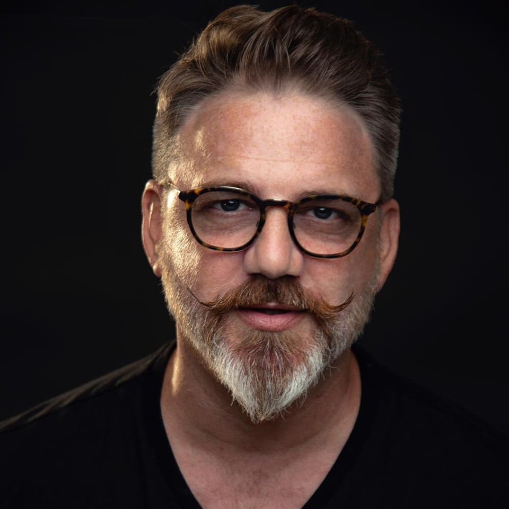 A color portrait of a middle-aged man with a meticulously groomed ducktail beard. His beard is full and tapers to a neat point at the chin, resembling the shape of a duck's tail. The beard is a mix of salt-and-pepper, with darker tones near the mustache and lighter, almost white hues at the chin. The man sports a styled mustache with curled tips, adding a touch of vintage flair to his modern look. He is wearing round, tortoiseshell glasses that complement his facial features. His hair is short on the sides and longer on top, brushed back neatly. He is dressed in a simple black shirt, which enhances the focus on his well-groomed beard and mustache. The background is dark, ensuring that the attention remains on his face and beard. The lighting is soft and focused, highlighting the texture and precision of his grooming, giving the portrait a professional and polished appearance.