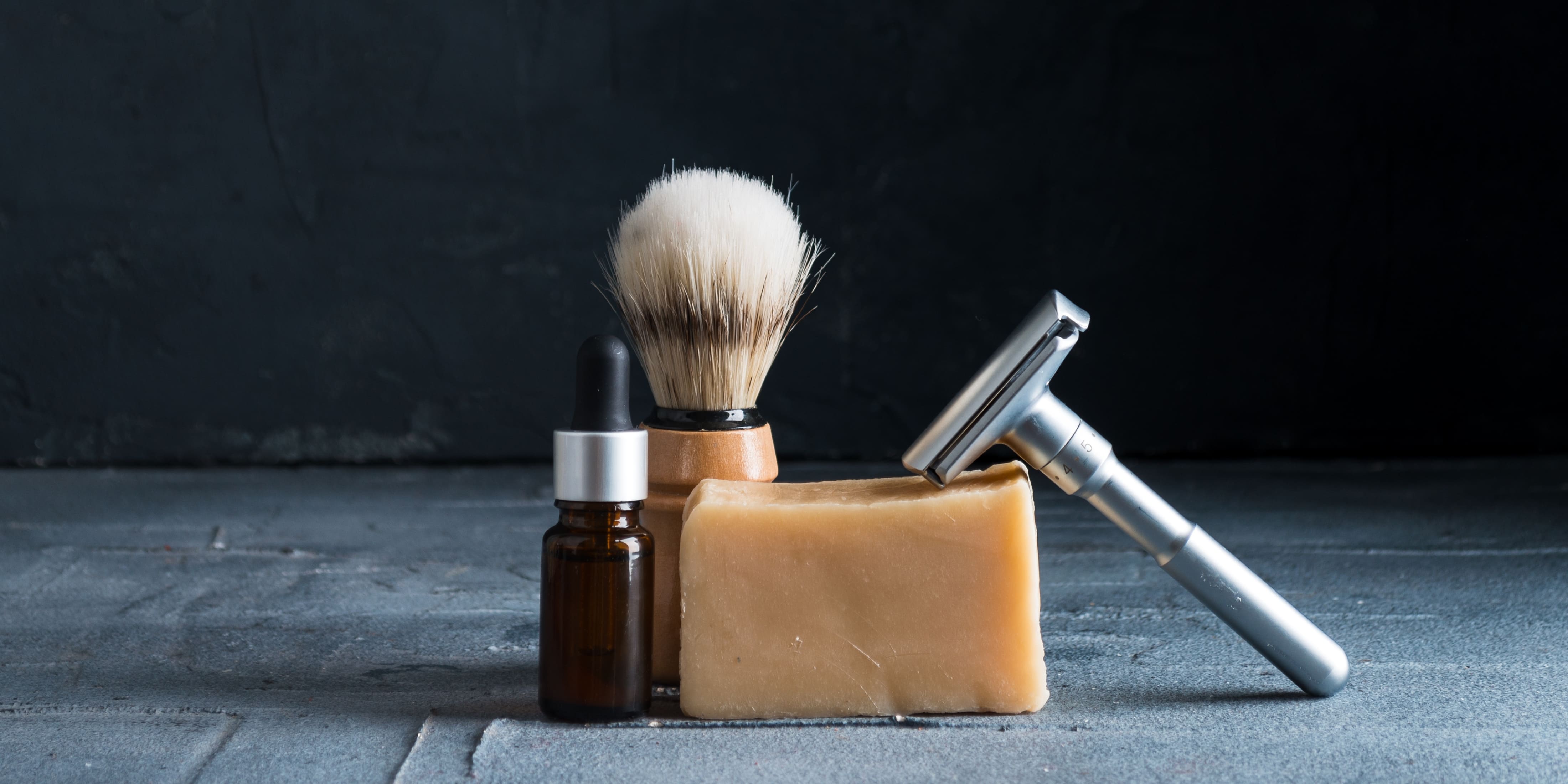 A collection of essential shaving tools for how to get a close shave, including a safety razor, shaving brush, bar of shaving soap, and a bottle of pre-shave oil. These items are neatly arranged against a dark, textured background, highlighting their importance in achieving a close shave.