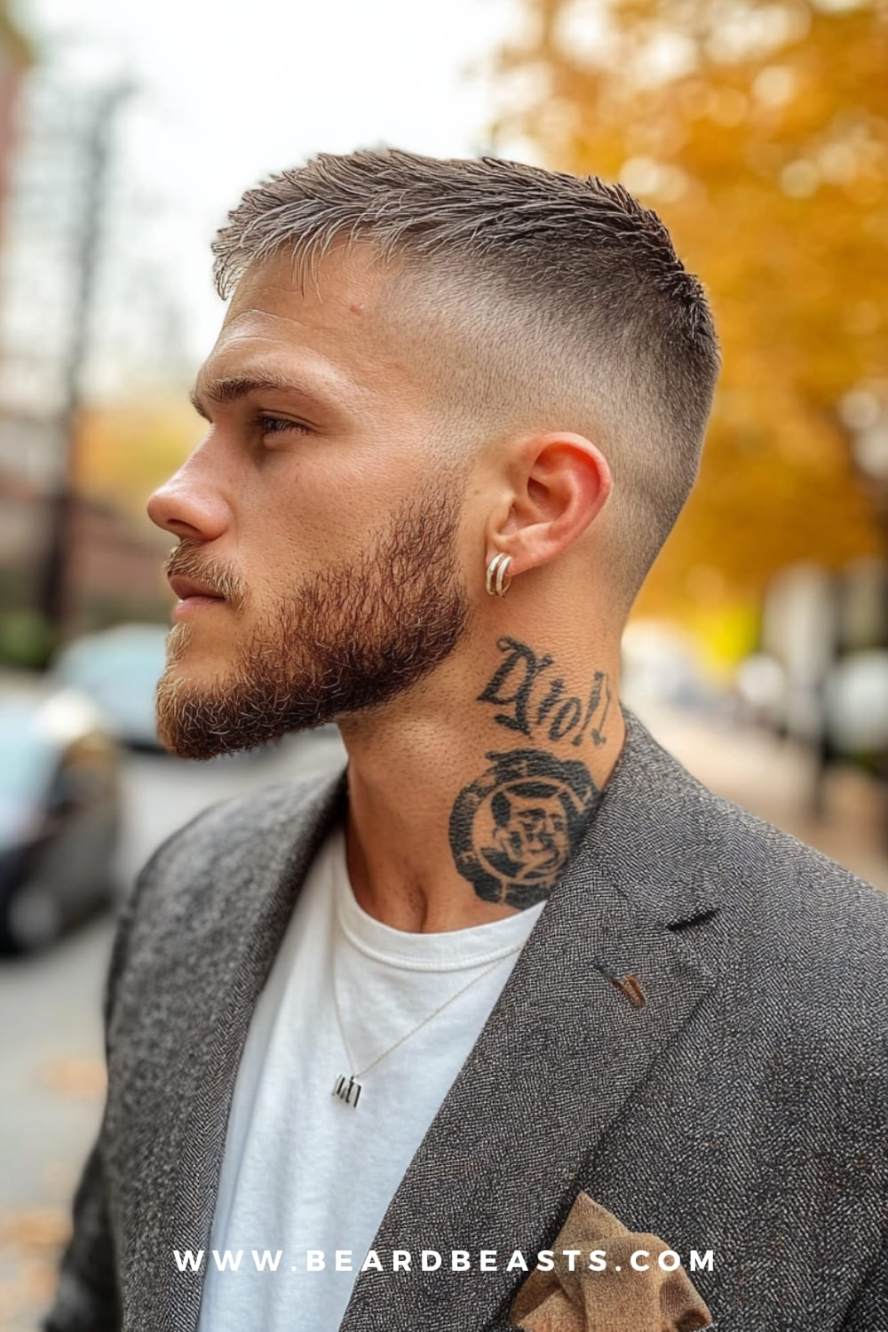 A man featuring a stylish fade haircut with a gradual transition from longer textured hair on top to shorter sides. The look is paired with a groomed beard and visible neck tattoos, showcasing a modern, edgy yet polished style.