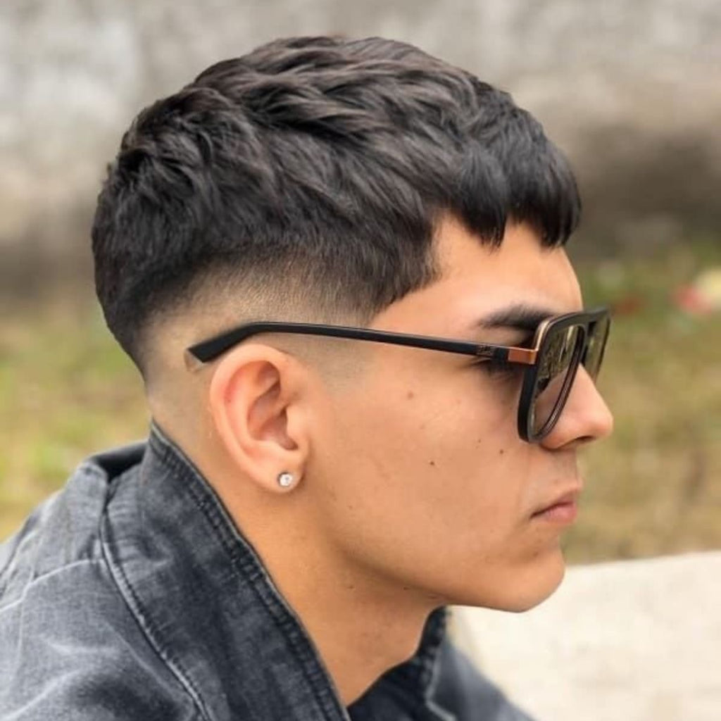 A side portrait of a young man wearing stylish black sunglasses, sporting a French crop mid fade haircut. The top of his hair is textured with short, choppy layers, while the mid fade on the sides gradually blends into the skin, creating a sharp and modern look. He is dressed in a casual denim jacket, accessorized with a small stud earring. The fade and textured top contrast nicely, adding to the overall edgy vibe. The outdoor background is blurred, keeping the focus on his trendy French crop mid fade and cool, confident style.