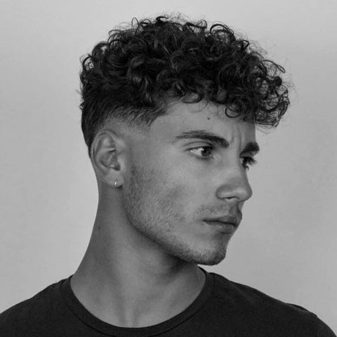 A young man is depicted with a curly fringe haircut, where his naturally curly hair is styled into a full, textured fringe that sits just above his forehead. The curls are dense and well-defined, giving the top of his head a voluminous, lively appearance. The sides are neatly faded, gradually blending into his skin, which contrasts with the thick curls on top. His expression is thoughtful, with his gaze directed slightly to the side, highlighting the strong lines of his jaw and cheekbones. He is wearing a simple, dark t-shirt, which keeps the focus on his hairstyle. The image is in black and white, emphasizing the texture and shape of the curls and the sharpness of the fade, creating a timeless and classic look. The overall style is youthful, clean, and modern, ideal for men embracing their natural curls.