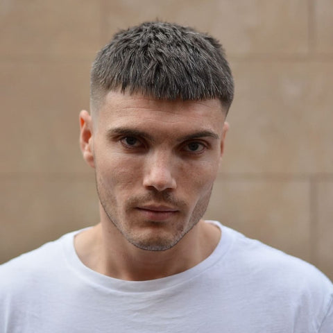 A young man is shown with a straight fringe haircut, featuring short, neatly trimmed bangs that rest just above his eyebrows. His hair is cut close to the scalp on the sides, creating a sharp fade that contrasts with the slightly longer, textured hair on top. The fringe is blunt and evenly cut, giving his hairstyle a clean and modern look. He has a serious, focused expression, which complements the precision and structure of his haircut. The man is dressed in a simple white t-shirt, enhancing the minimalist and clean aesthetic of his overall appearance. The background is a neutral-toned wall, which allows the details of his straight fringe and sharp fade to stand out clearly, emphasizing the crisp lines and well-groomed style.