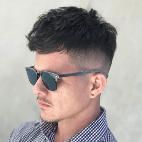 A young man is depicted with a stylish fringe haircut suited for thick hair. His dark, thick hair is cut into a textured fringe that sits neatly across his forehead, with the top left slightly tousled to add volume and movement. The sides feature a high fade, creating a clean and sharp contrast with the fuller hair on top. He is wearing sleek, dark sunglasses and a checkered button-up shirt, adding a touch of sophistication to his overall look. The man’s expression is calm and confident as he gazes off into the distance, embodying a modern and polished style. The background is a soft, neutral color, which helps to highlight the details of his haircut and fashion choices, making the thick, textured fringe the focal point of the image.