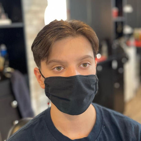 A young man is shown with a fringe hairstyle that is well-suited for thin hair. His hair is parted down the middle, with the fringe gently sweeping to each side, creating a soft and natural look that frames his face. The hair is slightly longer on top, providing volume and texture, while the sides are neatly trimmed for a clean finish. He is wearing a black face mask, which covers the lower half of his face, leaving his expressive eyes as the focal point. The man is dressed in a simple dark shirt, and the background appears to be a barbershop, with blurred details of barbering tools and equipment in the background. The overall look is neat and understated, perfect for those with thinner hair who want a low-maintenance yet stylish haircut.