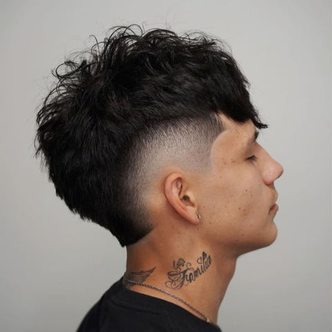 A young man is shown with a fringe haircut paired with a burst fade. His hair is thick and dark, with the top styled into a textured, slightly messy fringe that extends down to cover part of his forehead. The burst fade is prominently featured, with the hair around his ears gradually tapering down to the skin, creating a clean and sharp contrast between the longer top and the closely faded sides. The fade curves around his ears, accentuating the shape of his head and highlighting the volume of the hair on top. He has a small earring in his left ear and visible tattoos on his neck, adding to the edgy and modern aesthetic of his look. The background is plain, allowing the details of his hairstyle and personal style to stand out clearly. His expression is calm and composed, enhancing the overall cool and contemporary vibe of the image.