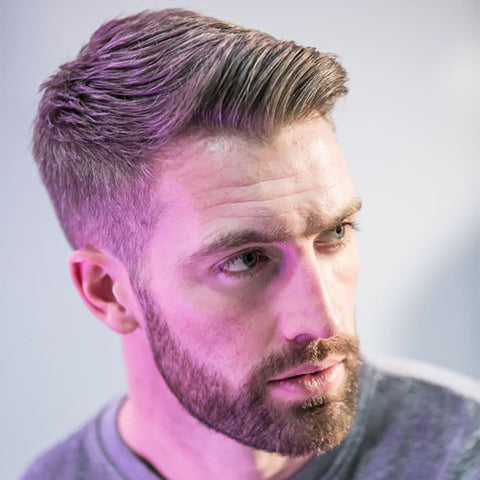 A man is shown with a fringe hairstyle that transitions into a quiff, creating a stylish and well-groomed appearance. His hair is light brown, with the top styled into a voluminous quiff that is swept back and slightly to the side, giving the hairstyle height and a refined shape. The sides are neatly trimmed, blending smoothly into the longer hair on top. He has a full beard that is carefully groomed, complementing the sleek hairstyle and adding to his masculine look. The lighting in the image casts a soft purple hue across his face and hair, adding a modern and slightly artistic touch to the photograph. The man’s expression is thoughtful, with his gaze directed slightly downward, further emphasizing the sophisticated and polished nature of his appearance. The background is blurred, keeping the focus on his quiff hairstyle and well-maintained beard, making this look ideal for men who appreciate classic styles with a contemporary twist.