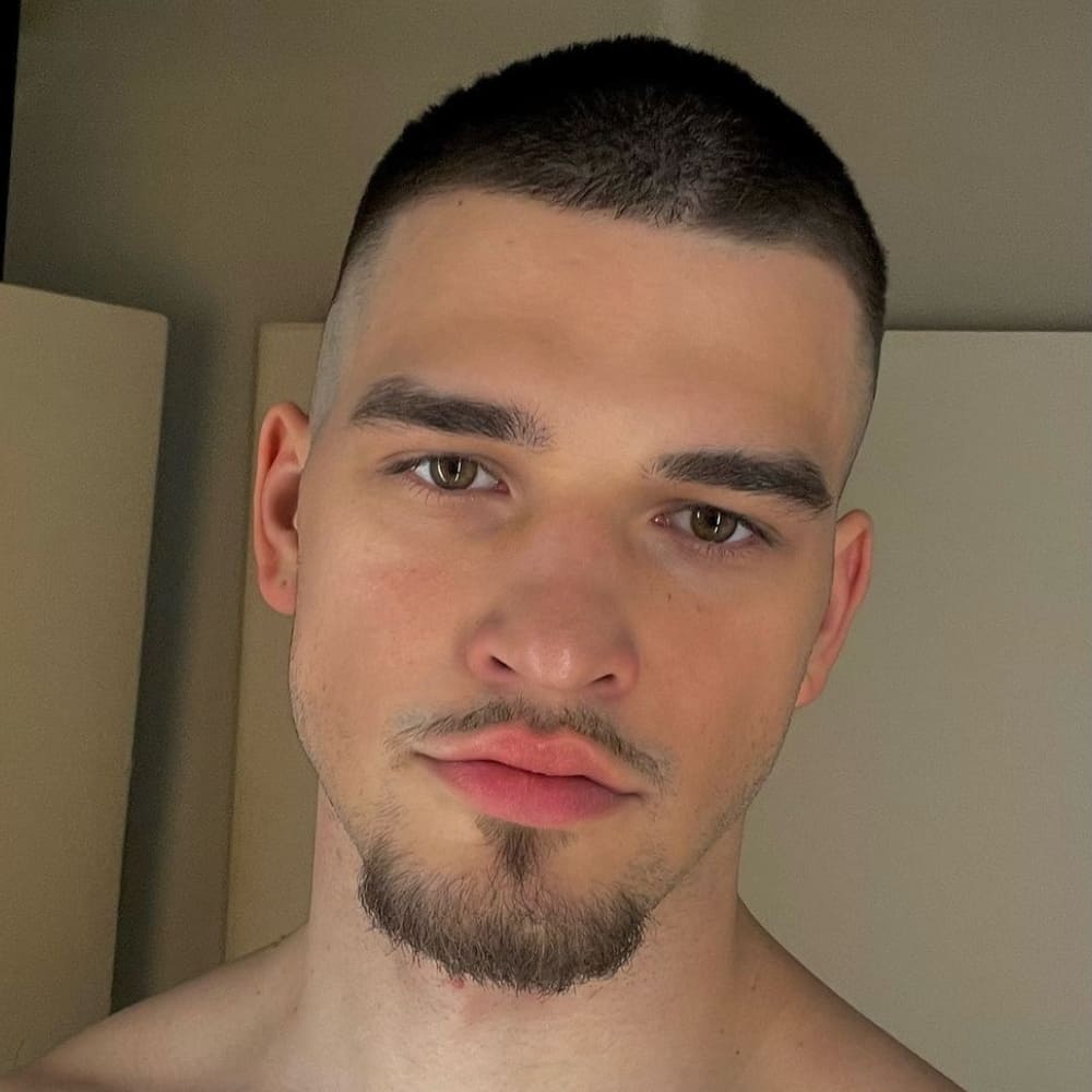 A close-up portrait of a young man with a neatly styled goatee beard. His facial hair is focused on the chin and connected to a mustache, creating a distinct goatee look. The goatee is well-groomed, highlighting his strong chin and jawline. The man has short, buzz-cut hair that complements the clean and sharp appearance of his beard. He has clear, light-colored eyes and a neutral expression, with his lips slightly parted. The background is simple and uncluttered, putting emphasis on his facial features and beard style. The lighting is soft, enhancing the natural tones of his skin and the precision of his grooming.