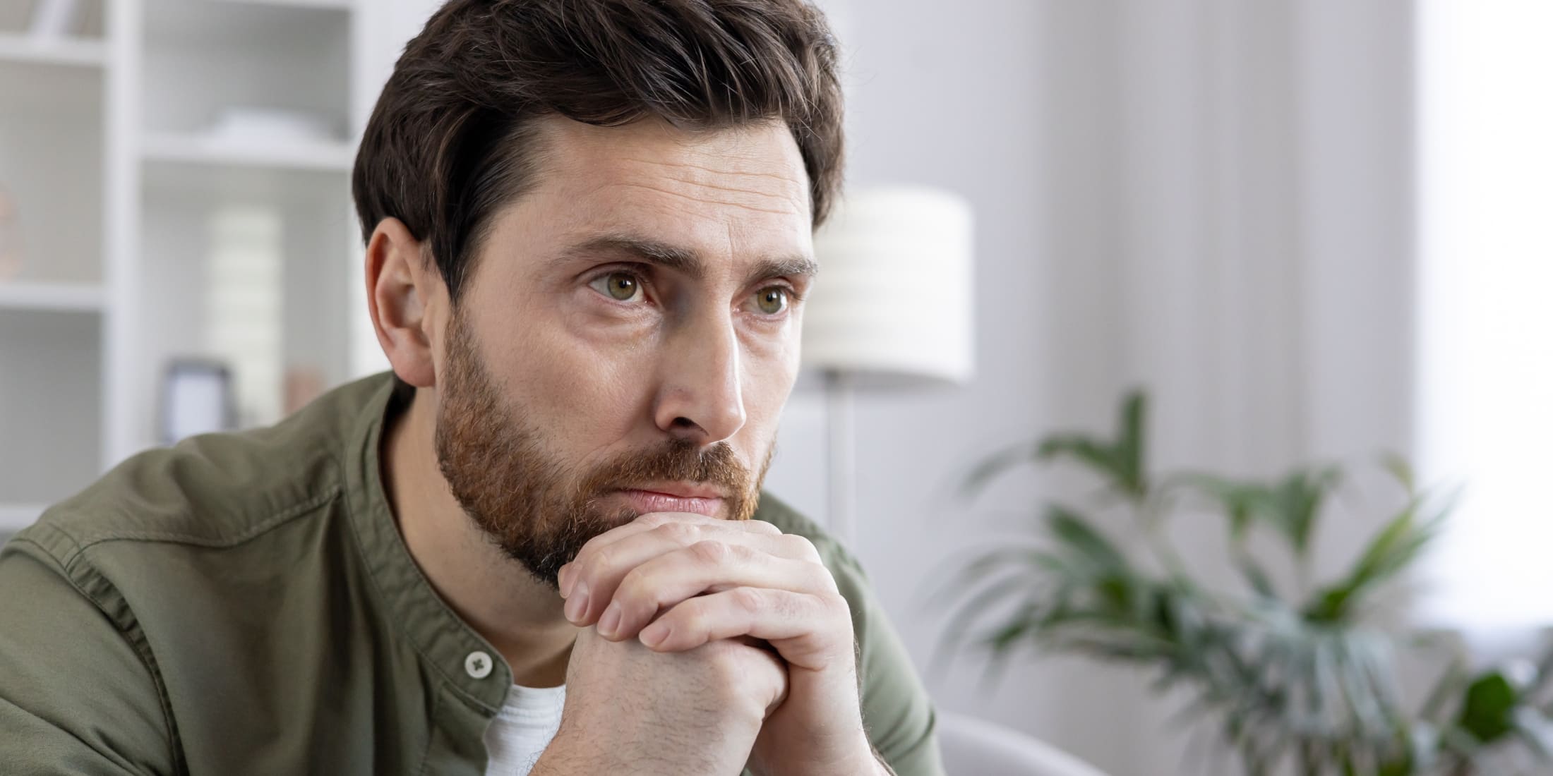 Man with a contemplative expression, resting his chin on his hands while sitting indoors. The image conveys a moment of deep thought, reflection, or concern, highlighting the importance of mental health habits for men, such as mindfulness and stress management. The calm, neutral background with indoor plants adds to the serene and introspective atmosphere.