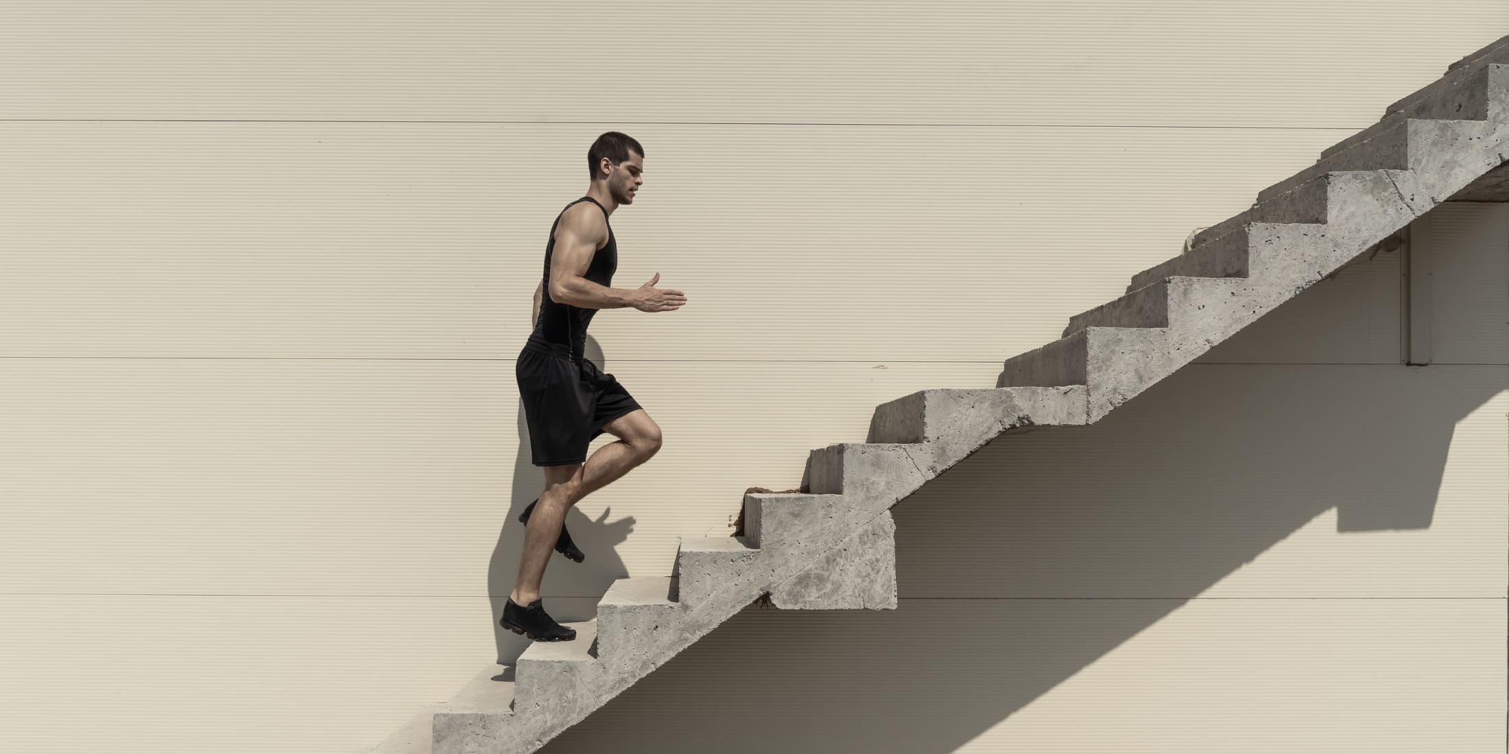 Man in athletic attire running up a set of outdoor concrete stairs, symbolizing progress and determination. The image emphasizes personal growth, illustrating the importance of consistent effort, self-discipline, and perseverance in achieving goals. The minimalist background draws attention to the man's focused upward movement, representing the journey toward personal and professional development.