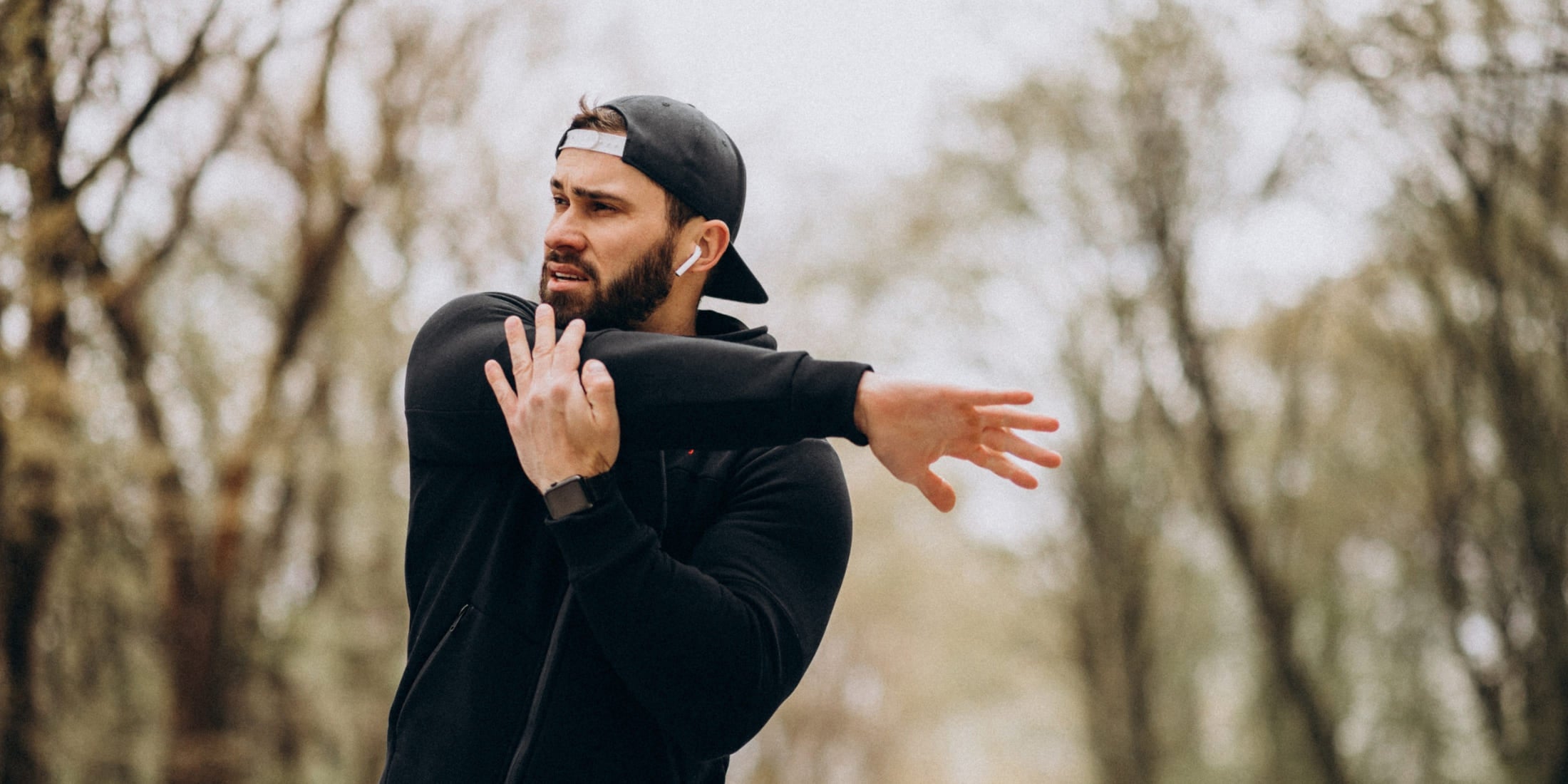 Man in athletic gear, wearing a cap and wireless earbuds, stretching his arm outdoors before a workout. This image captures the essence of habits for men focusing on physical health, emphasizing the importance of regular exercise, preparation, and staying active to maintain a healthy lifestyle. The natural background with trees suggests a serene environment ideal for fitness and well-being.