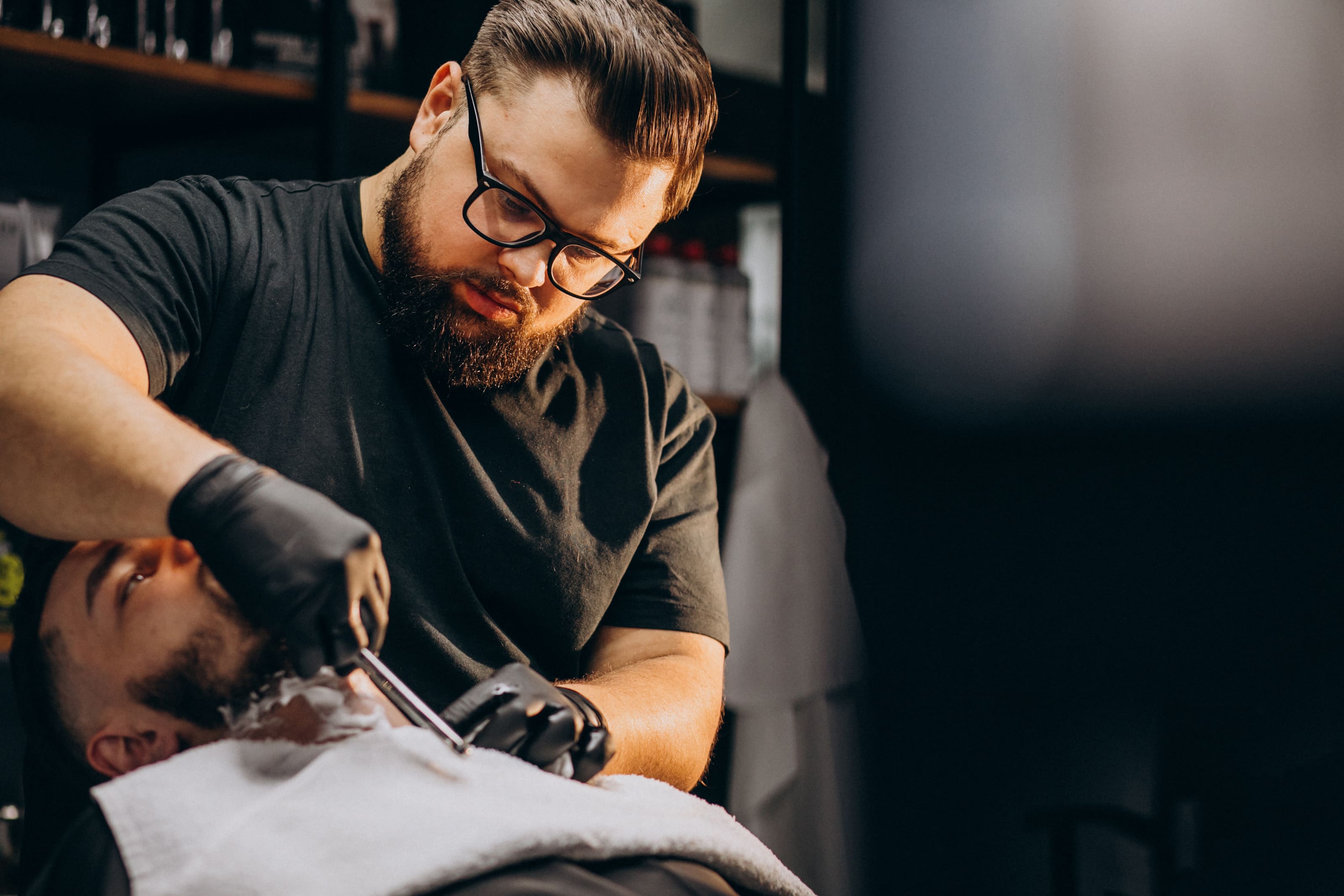 A bearded barber in black gloves and a black T-shirt is carefully shaping another man's beard with an electric trimmer. The customer, reclined in a barber chair with a towel around his neck, appears relaxed. The setting suggests a modern barbershop with product shelves visible in the background.