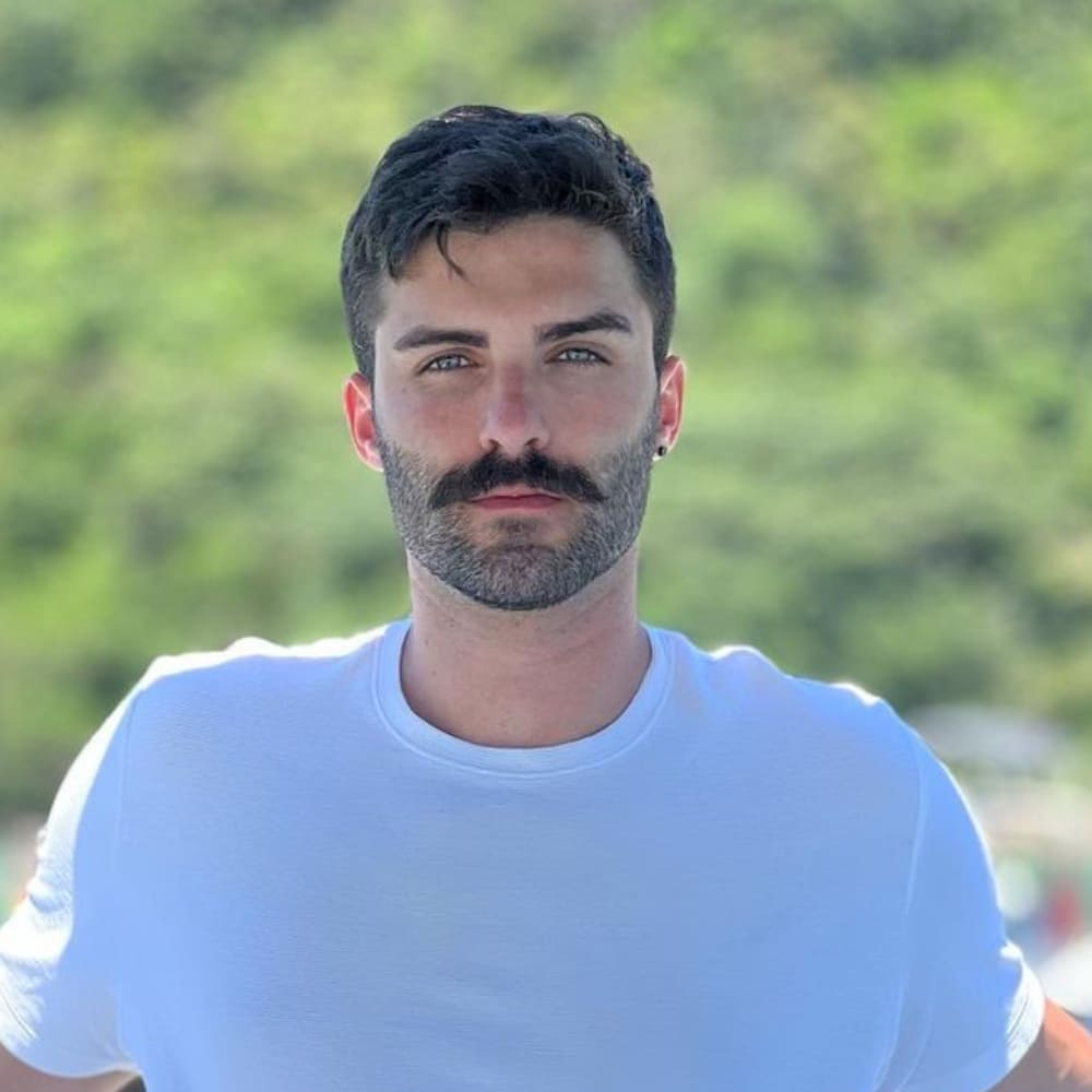 A man showcasing a heavy stubble beardstache, characterized by a thick, well-defined mustache and heavy stubble beard. He has short, dark hair and is wearing a white t-shirt. The background features a lush, green outdoor setting, highlighting the natural and masculine appeal of the heavy stubble beardstache style. The overall look is rugged and stylish, perfectly capturing the essence of this facial hair trend.