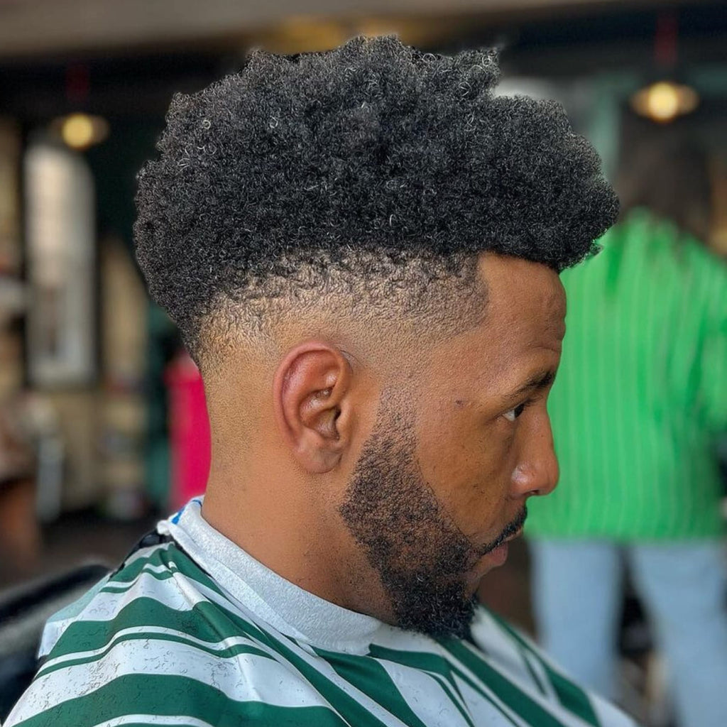 Side profile of a man with a high-top haircut featuring a sharp, clean taper along the sides and back. The voluminous natural texture on top contrasts beautifully with the neatly tapered sides, creating a bold and stylish look. The beard is shaped and blended seamlessly with the haircut, adding to the overall polished appearance. A modern yet classic style that emphasizes natural curls and sharp lines.