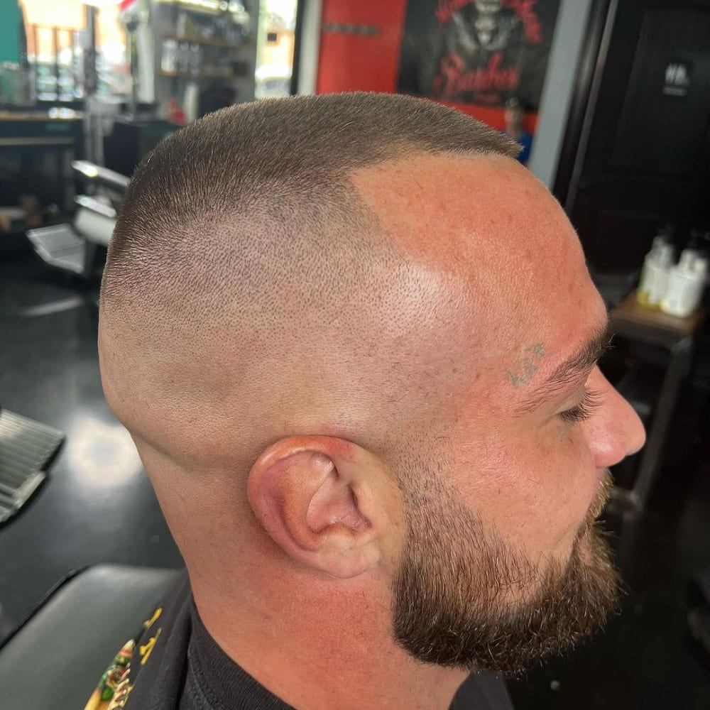 A side profile of a man with a high and tight buzz cut. The haircut features a very short length on top that seamlessly transitions into a close-shaved, almost bald fade on the sides and back. The high and tight style emphasizes the sharp contrast between the top and the sides, creating a bold, clean look. The man also has a neatly trimmed beard that complements the angular and defined lines of the haircut, giving a strong and polished appearance. The overall style is modern and military-inspired, highlighting the crisp edges and the neat, minimalist finish.