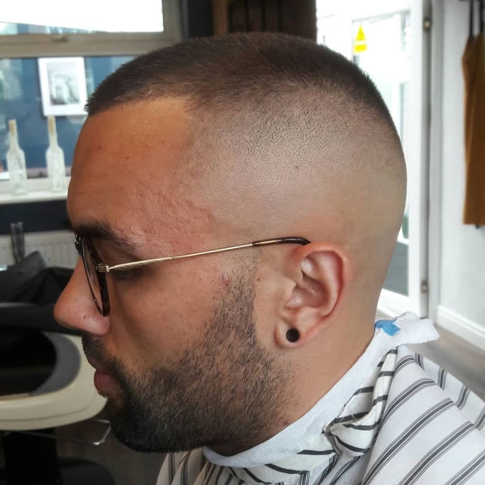 The image shows a person with a high and tight buzz cut haircut. The sides and back of the head are shaved very close to the skin, creating a high fade. The top of the head is uniformly short, closely cropped to match the length of the sides and back, giving it a clean, minimalist look. The overall style is practical and low maintenance, ideal for those who prefer a straightforward, no-fuss haircut. The person is also wearing glasses and has a neatly trimmed beard, adding to the clean and sharp appearance.