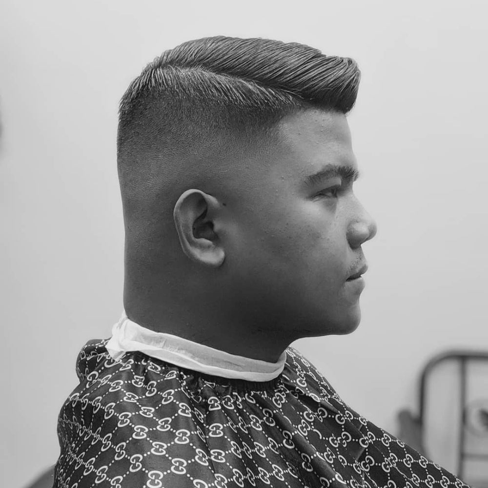 The image shows a person with a high and tight fade haircut. The sides and back of the head feature a smooth, gradual fade from very short at the bottom to slightly longer as it moves up towards the top. The top of the head has longer hair, styled neatly with a slight wave and a side part. This style blends the classic high and tight with a modern fade, creating a seamless transition between the lengths. The overall appearance is contemporary and stylish, suitable for various settings, from professional to casual. The person’s hair is meticulously groomed, showcasing the clean lines and precise fade.