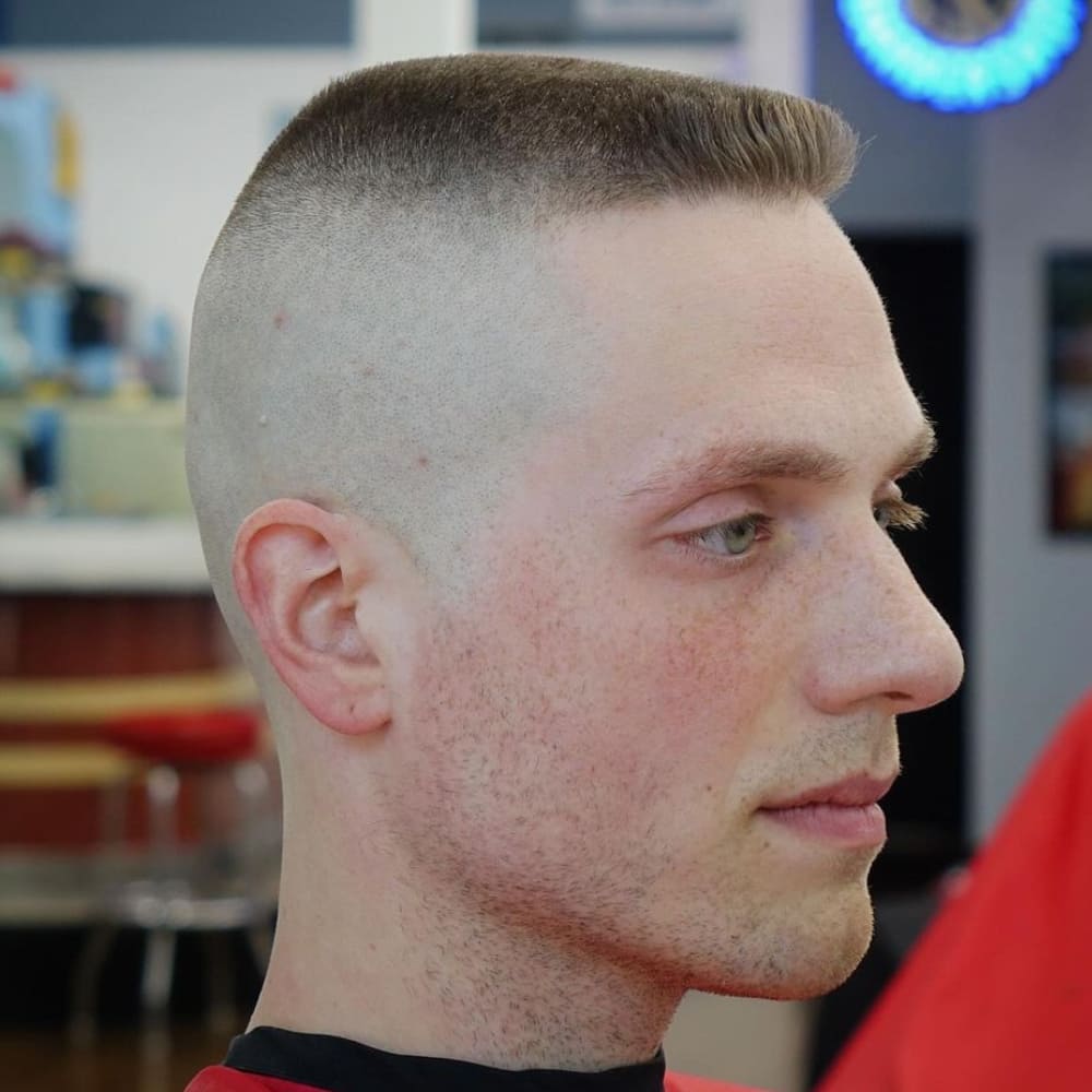 The image shows a person with a high and tight flat top haircut. The sides and back of the head are shaved very close to the skin, featuring a high fade. The top of the head is cut flat and level, creating a distinctive boxy shape that stands out. The flat top is neatly trimmed and styled to maintain its straight, horizontal appearance. This hairstyle combines the clean lines of a high and tight with the retro, edgy vibe of a flat