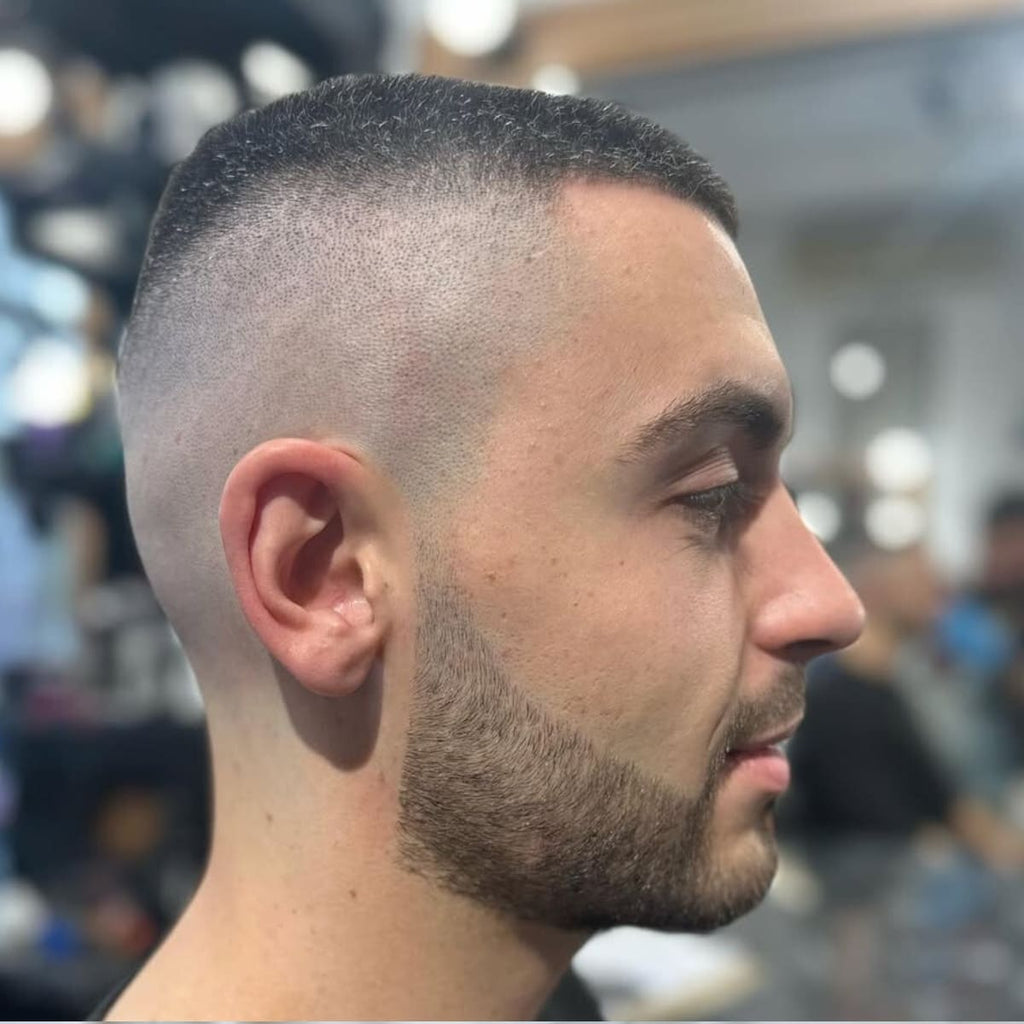 A side profile of a man with a skin fade buzz cut, featuring a sharp high fade that transitions smoothly from the skin to short, textured hair on top. The haircut is paired with a neatly groomed beard, blending seamlessly into the fade for a clean and polished look. The precise lines and bold fade highlight the modern and edgy appeal of this low-maintenance style.