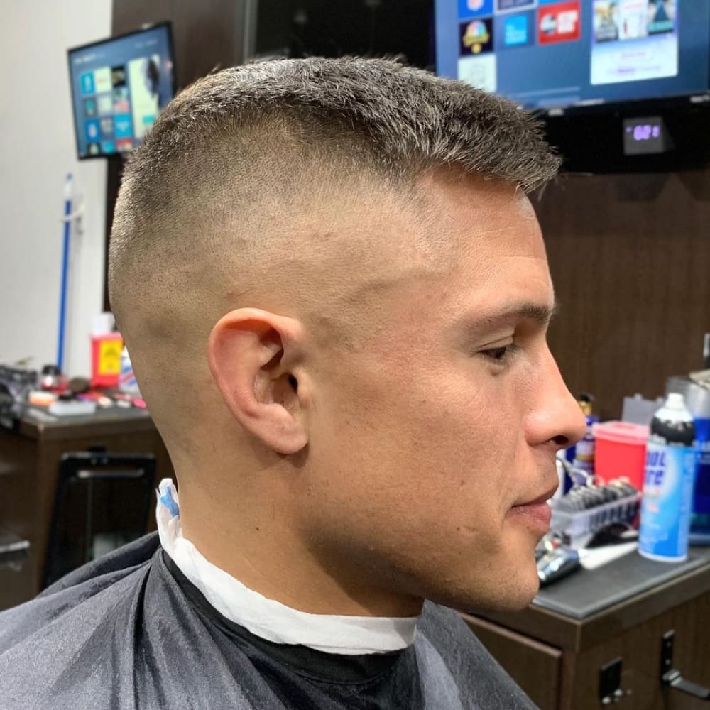 The image shows a person with a high and tight skin fade haircut. The sides and back of the head are shaved down to the skin, creating a very clean and sharp high fade. The top hair is left slightly longer, around an inch or less, maintaining a neat and tidy appearance. This ultra-clean fade offers a polished and contemporary look. The person is seated in a barbershop, with grooming tools and products visible in the background, emphasizing the fresh and precise styling of the haircut. The overall appearance is modern, professional, and easy to maintain.