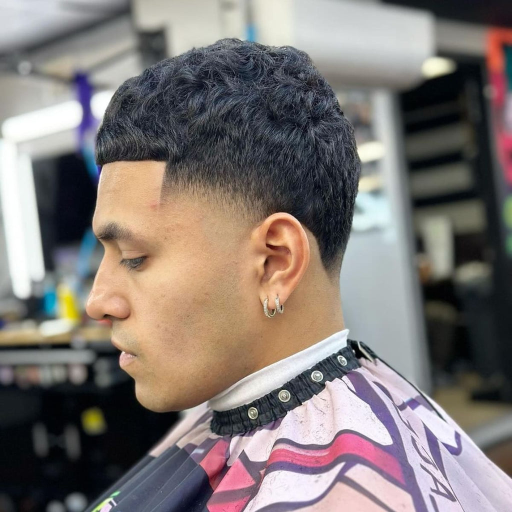 Profile view of a man sporting a high bald taper fade haircut, featuring a sharp fade starting high on the head and tapering down smoothly into the skin. The style complements the textured, curly top, creating a striking contrast between the faded sides and fuller crown. Perfect for a modern, edgy look, this high bald taper fade adds a clean, structured element to curly hair while maintaining a sharp, defined hairline.