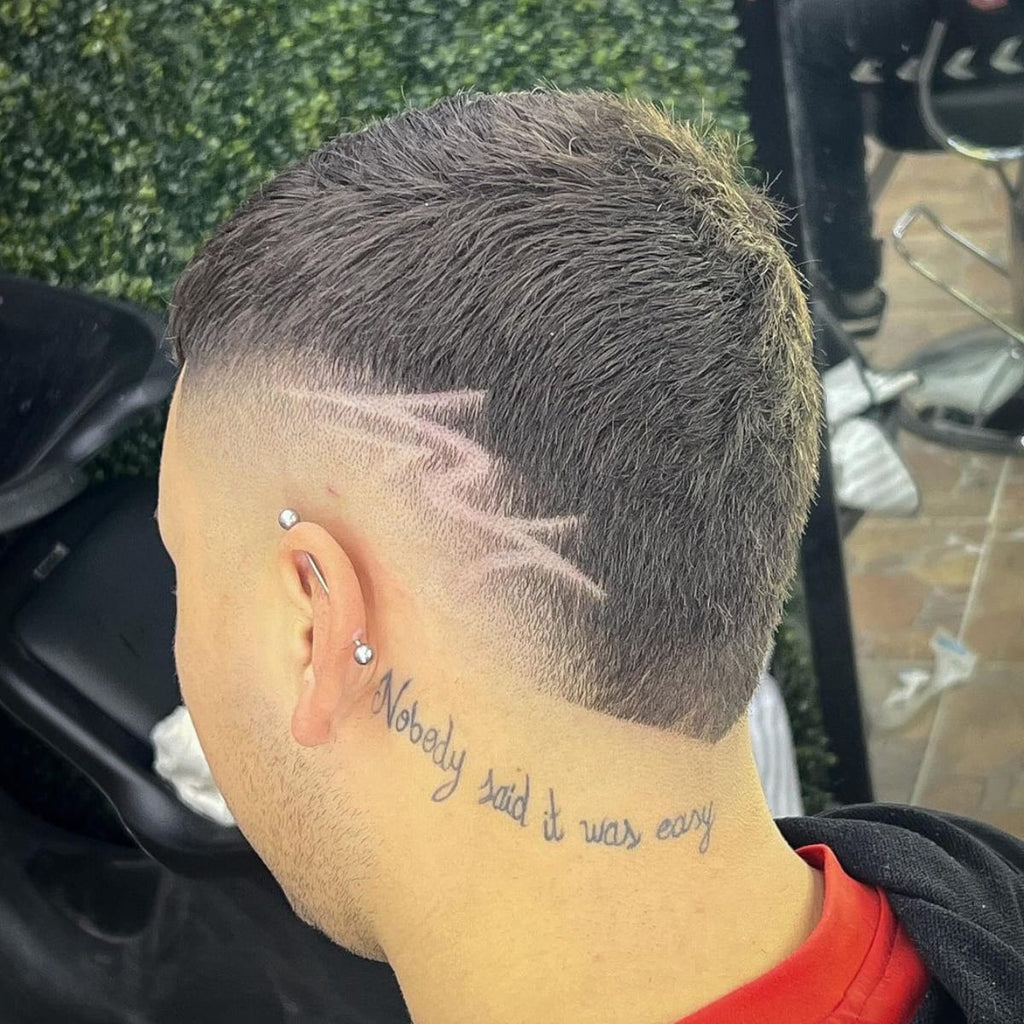 Back view of a man with a high burst fade featuring a sharp lightning bolt design shaved into the fade. The edgy hairstyle is complemented by a neck tattoo that reads 'Nobody said it was easy' and a stylish ear piercing, giving the look a bold, creative edge.