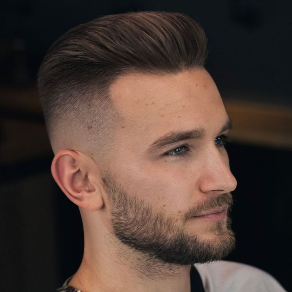 A man is showcasing a modern pompadour haircut with a high fade. His hair on top is neatly styled upwards and backwards, creating a sleek and voluminous look. The sides are sharply faded, enhancing the contrast between the full top and the short sides. His beard is well-groomed, adding a touch of ruggedness that complements the sophisticated hairstyle. The overall effect is a contemporary and stylish appearance, perfect for 2024 trends.