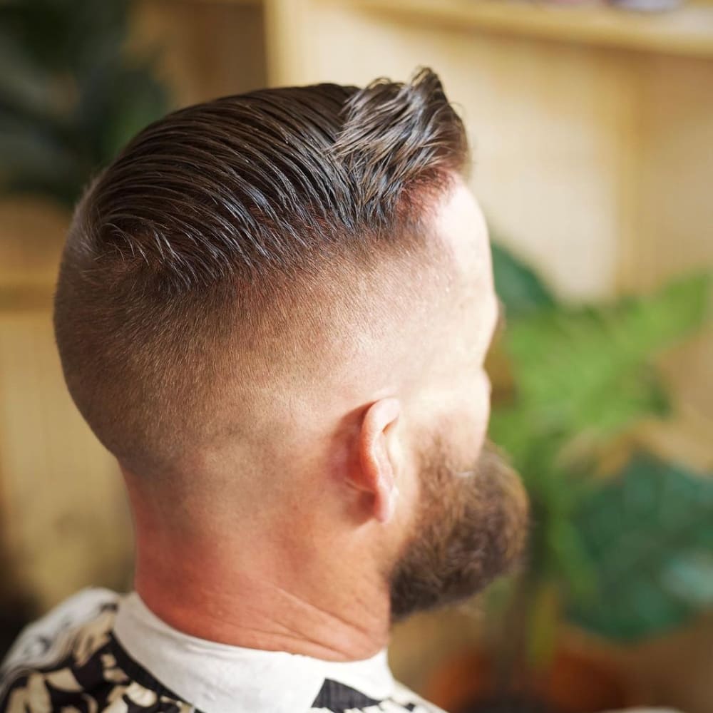 A man with a meticulously styled high fade and comb over haircut. The hair on top is smoothly combed over to one side, creating a sleek and polished appearance. The high fade begins just above the temples and transitions sharply down to the skin, emphasizing the clean, precise lines of the fade. The man has a well-groomed beard that complements the hairstyle, adding to the overall sharp and sophisticated look. The background features a blurred, indoor setting with some greenery, keeping the focus on the stylish haircut.