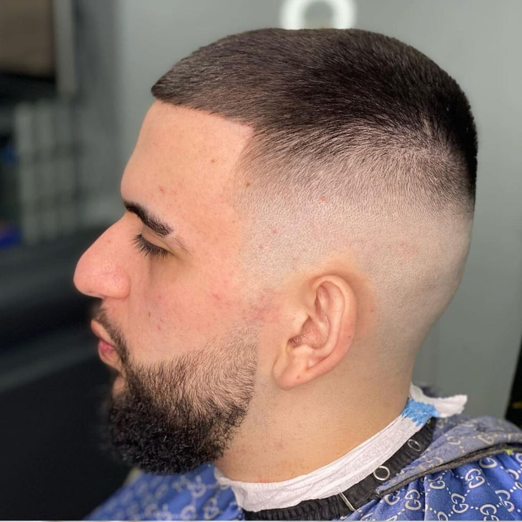 A side profile of a man featuring a clean skin fade buzz cut, with the fade transitioning smoothly from bare skin to a short, even length on top. The hairstyle is complemented by a neatly shaped full beard, creating a balanced and polished look. The sharp fade highlights the precise edges and enhances the modern, professional vibe of the cut.