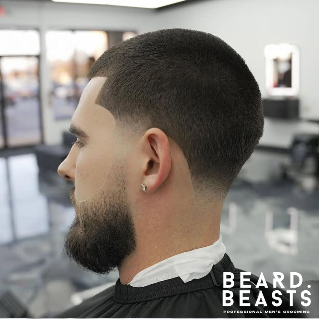 A side profile of a man with a high taper fade buzz cut, featuring a clean tapering effect from skin at the nape to short, uniform hair on top. The haircut is accompanied by a neatly groomed full beard, blending seamlessly with the taper for a sharp, polished look. A defined lineup adds extra precision to the style, while the taper creates a soft, gradual transition. The background showcases a bright, modern barbershop with glossy floors and natural light, emphasizing the professional grooming quality. The 'Beard Beasts' logo is visible in the corner, symbolizing expert men's grooming.