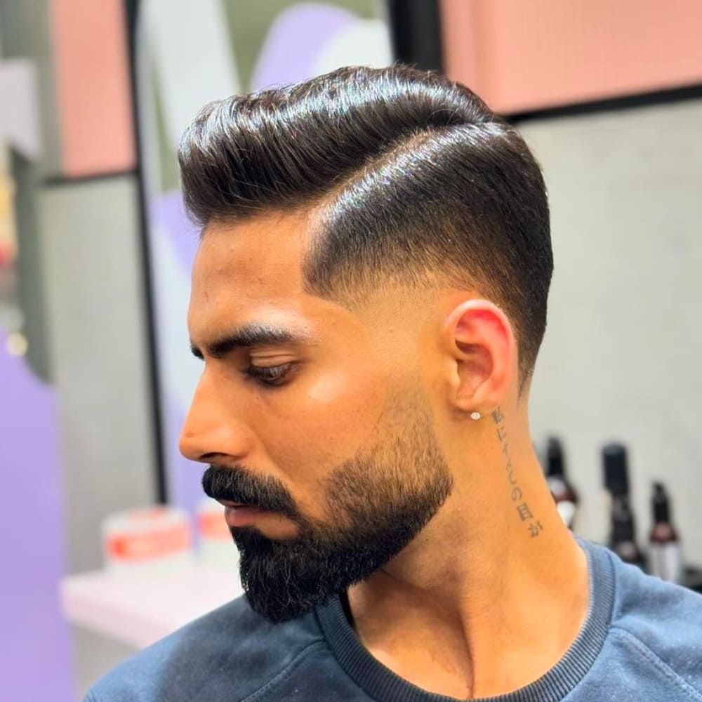 Man with a stylish high taper side part haircut, featuring clean lines, a well-defined part, and neatly tapered sides and back, complemented by a full beard and a tattoo on his neck.